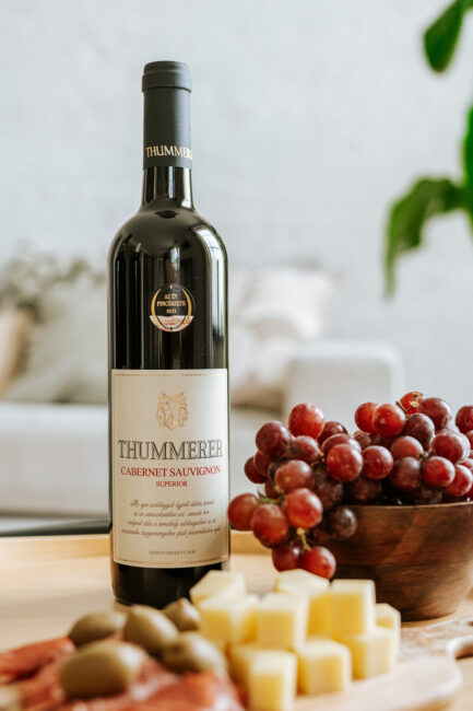 Egy palack Egri Cabernet Sauvignon Superior 2021 bor szőlőtál és sajttál között.